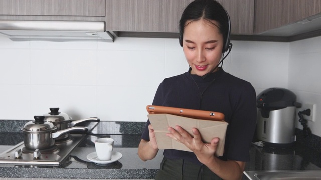 亚洲女性戴着耳机和使用数字平板电脑视频会议和会议，在家里的厨房喝咖啡