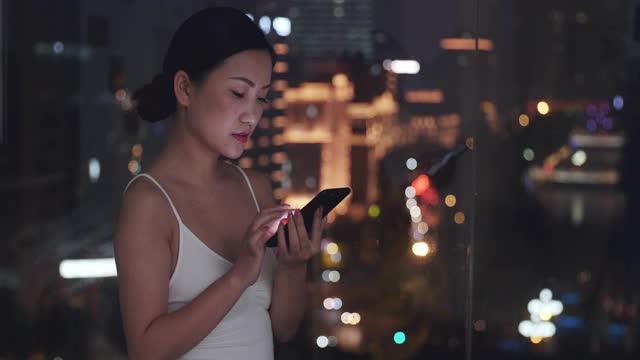 年轻漂亮的亚洲女人晚上在窗边用手机