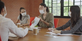 一群年轻的亚洲创意或商业人士戴着防护面具，在办公室一起开会