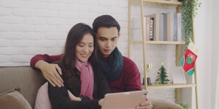 年轻的亚洲夫妇情人视频通话与平板电脑在圣诞节和快乐的新年假期节日在家。圣诞节庆祝活动的概念。4 k慢动作。