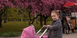 在公园里，一个小女孩推着一辆婴儿车走在前面，旁边是一棵开花的树。
