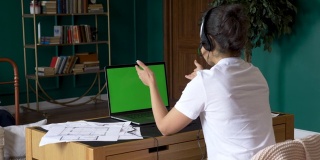 坐在椅子上的年轻女子在一台绿色模拟屏幕的笔记本电脑上工作。使用电脑的女孩，在线电话会议，与人培训，使用电脑。家中明亮的房间