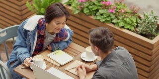 跟踪顶视图的年轻亚洲女性和白人男子讨论大学项目或创业的想法，坐在户外的咖啡桌上用笔记本电脑