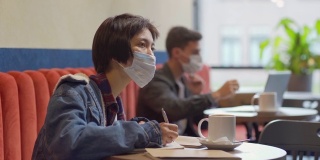 镜头从在笔记本电脑上工作的年轻白人男子，转向坐在咖啡馆桌旁浏览笔记本、做笔记和思考的亚洲女性，两人都戴着口罩
