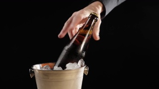 新鲜的啤酒。Hand将一个装有美味精酿啤酒的棕色瓶子放入一个装有冰的桶中，以黑色为背景。冰镇新鲜啤酒与水滴。准备饮料。4 k视频素材模板下载
