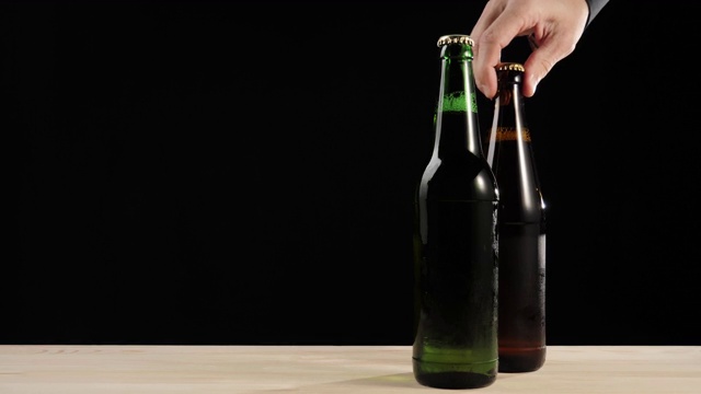 新鲜的啤酒。Hand把一个装有美味精酿啤酒的棕色瓶子和一个装有啤酒的绿色瓶子放在一张黑色背景的木桌上。冰镇新鲜啤酒与水滴。准备饮料。广角镜头。4 k