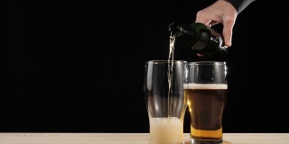 新鲜的啤酒。手从一个棕色的瓶子里将美味的精酿啤酒倒在啤酒杯架上的第二个杯子里，倒进一张黑色背景的木桌里。冰凉的新鲜啤酒，带有水滴，泡沫和泡沫。4 k