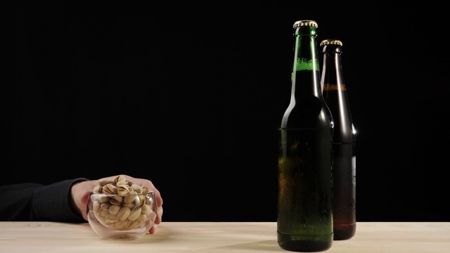 新鲜的啤酒。Hand拿着一碟开心果，旁边是绿色和棕色的瓶子，上面放着美味的精酿啤酒，放在一张黑色背景的木桌上。冰镇新鲜啤酒与水滴。4 k
