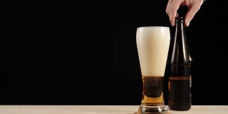新鲜的啤酒。Hand把一个棕色的瓶子和一瓶美味的精酿啤酒放在一张黑色背景的木桌上。冰凉的新鲜啤酒，带有水滴，泡沫和泡沫。准备饮料。4 k