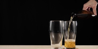 新鲜的啤酒。在黑色背景的木桌上，手从一个棕色的瓶子里将美味的精酿啤酒倒进啤酒杯。冰凉的新鲜啤酒，带有水滴，泡沫和泡沫。4 k
