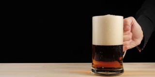 新鲜的啤酒。手把一杯美味的手工啤酒放在啤酒杯里，放在一张黑色背景的木桌上。冰凉的新鲜啤酒，带有水滴，泡沫和泡沫。4 k