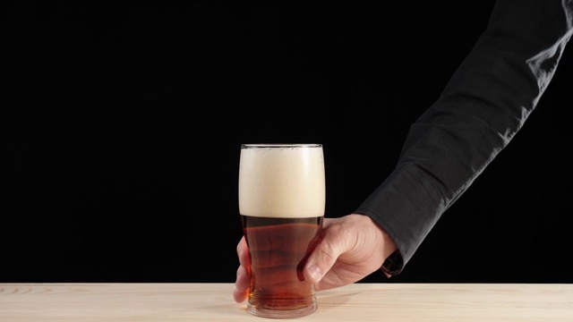 新鲜的啤酒。Hand从一张黑色背景的木桌上拿起一只装在啤酒杯里的美味精酿啤酒。冰凉的新鲜啤酒，带有水滴，泡沫和泡沫。广角镜头。4 k