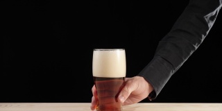 新鲜的啤酒。Hand从一张黑色背景的木桌上拿起一只装在啤酒杯里的美味精酿啤酒。冰凉的新鲜啤酒，带有水滴，泡沫和泡沫。广角镜头。4 k