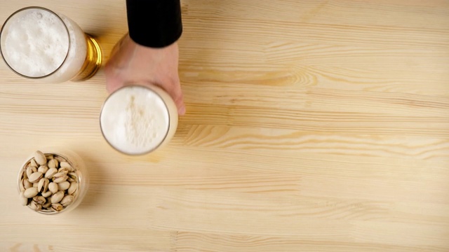 新鲜的啤酒。Hand从背景的木桌上拿了一杯装着开心果的玻璃杯和碟子，旁边放着另一个杯子。冰凉的新鲜啤酒，带有水滴，泡沫和泡沫。前视图。4 k