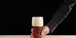 新鲜的啤酒。Hand把一杯装在啤酒杯里的美味精酿啤酒放在一张黑色背景的木桌上。冰凉的新鲜啤酒，带有水滴，泡沫和泡沫。广角镜头。4 k