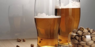 新鲜的啤酒。白色背景的木桌上放着两杯美味的冰啤酒，旁边放着一碟开心果。冰凉的新鲜啤酒，带有水滴，泡沫和泡沫。中间。4 k