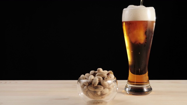 新鲜的啤酒。美味的精酿啤酒放在啤酒杯里，旁边放着一碟开心果，放在一张黑色背景的木桌上。冰凉的新鲜啤酒，带有水滴，泡沫和泡沫。广角镜头。4 k