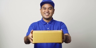 微笑年轻的亚洲快递员在蓝色制服给包裹硬纸板客户在孤立的白色背景。4 k决议。