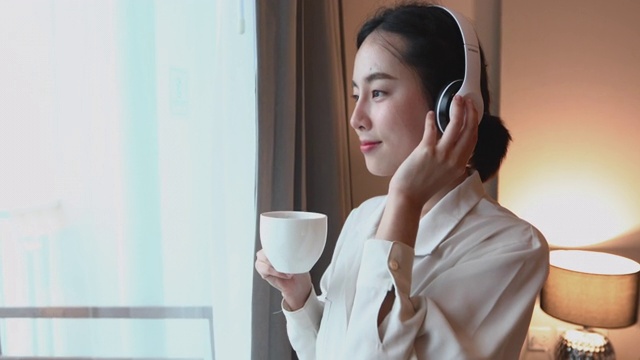 亚洲女性喜欢戴着耳机在手机上听音乐，在家里放松的时候喝咖啡