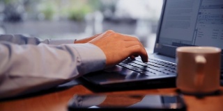 使用笔记本电脑工作的商务人士。穿蓝色衬衫在键盘上打字的人。一个人在笔记本电脑上工作的特写。一个人在办公桌上的特写，笔记本电脑，咖啡杯和智能手机。