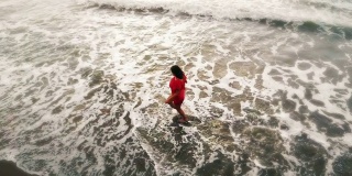 在日落的海边沙滩上，穿着红色裙子的女人在水里奔跑，鼓舞人心的自由快乐假日概念，从无人机空中拍摄，从上面看