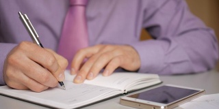 一个穿着紫色衬衫打着领带的商人在笔记本上做笔记。男人每天都写一支笔。一个人在记事本上做笔记。在办公室的办公桌上放笔记本电脑和智能手机。特写镜头。