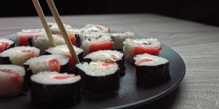 在盛有日本食物的盘子上轻轻敲击。女人用筷子从盘子里拿起寿司。寿司卷日本食物旋转在黑色的背景。加州寿司卷配金枪鱼，蔬菜，飞鱼卵和鱼子酱近距离旋转。日本