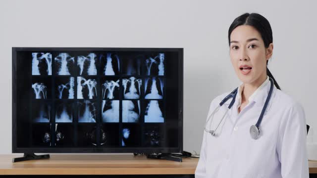 一名身穿实验服的亚洲女性，一边对着摄像机，一边在屏幕上解释x光胶片。