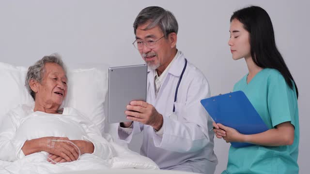 一位资深男医生拿着平板电脑向一位躺在医院病床上的资深女病人展示，一位女医生拿着写字板。