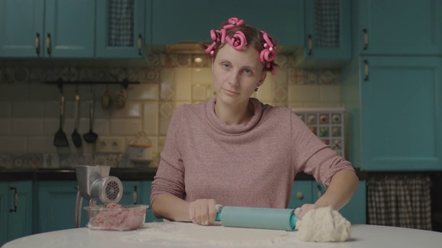 一名30多岁的年轻女子正用擀面杖擀面团，坐在厨房里看着摄像机。带卷发器在家独自做饭的家庭主妇。