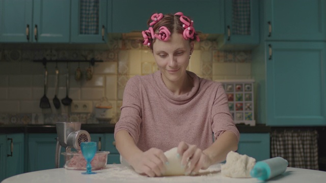 年轻的成年女子坐在厨房里在面团上挖洞。微笑的家庭主妇，带着卷发器独自在家做饭。面团滑稽的面具。