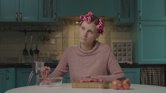 疲惫的30多岁的年轻女子，头上戴着卷发器，坐在厨房的绞肉机里绞肉。不快乐的家庭主妇独自在家做饭。