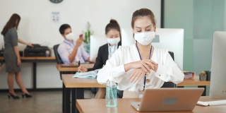 在新常态的办公室里，亚洲职场女性佩戴防护口罩、使用酒精凝胶洗手液以保持卫生，并保持社交距离，以防止冠状病毒新冠病毒的传播。
