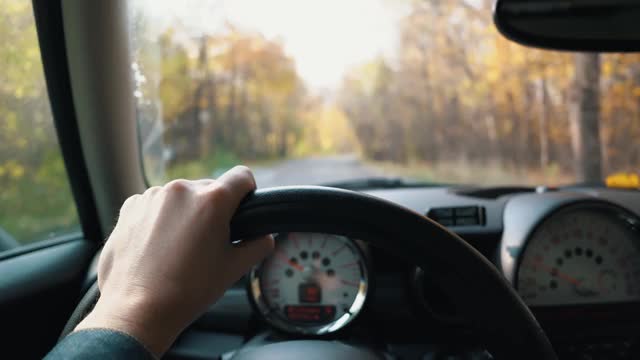 一个男人的手在方向盘上，在一个秋天的一天驾驶汽车。第一人称视角