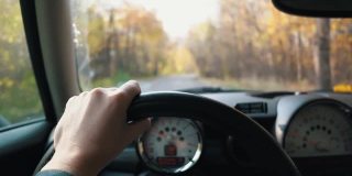 一个男人的手在方向盘上，在一个秋天的一天驾驶汽车。第一人称视角