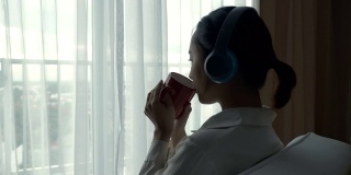 亚洲妇女放松听音乐与她的杯子靠近窗户的场景