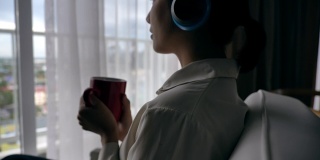 慢动作场景的亚洲女人放松听音乐靠近窗户与她的杯子
