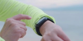 近距离观察亚洲健身女性的手在智能手表上设置健康跟踪，并在早上在海滩上慢跑时检查心率