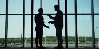 两个商人的剪影，握手和交谈站在办公大厅的大全景窗口，同事在慢镜头走过他们