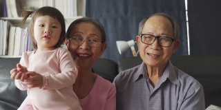 亚洲家庭在客厅里，爷爷奶奶和小外甥女孩对着镜头笑着玩耍。幸福享受居家生活。奶奶抱着可爱可爱的小孩宝宝坐在沙发上。