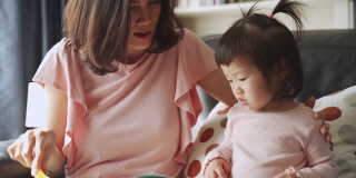 亚洲年轻美丽的母亲和她的小女儿一起在家里的沙发上看纪实书。他们指着书，看着它与幸福共处的时间。幸福的家庭的概念