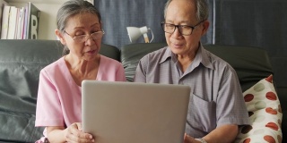 亚洲老夫妇，爷爷和奶奶在家里客厅的沙发上用笔记本电脑上网。老年人学习在生活方式活动中使用新科技。