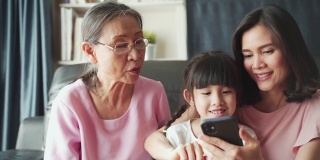亚洲家庭的孩子、母亲、祖母在客厅里用智能手机一起玩互联网，带着幸福和微笑。快乐活动科技生活方式手机使用概念。