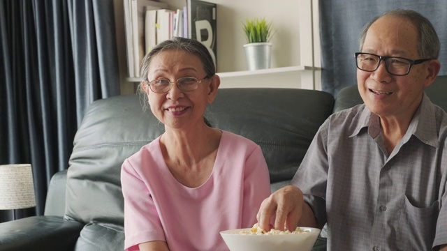 亚洲祖父母一起在家看电影。老人微笑着用遥控器看电视，笑得很开心。家庭中退休人员的休闲时间和生活方式活动。