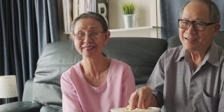 亚洲祖父母一起在家看电影。老人微笑着用遥控器看电视，笑得很开心。家庭中退休人员的休闲时间和生活方式活动。