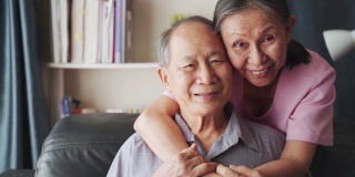 亚洲成熟夫妇坐在客厅微笑的肖像。老婆婆从后面抱住男人，幸福地看着镜头。退休后生活幸福，爷爷奶奶一起享受活动。