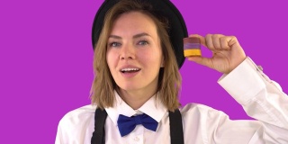 女商人展示扑克筹码，眨了眨眼睛，她戴着一顶黑帽子，领结，背带，袖扣和一件白衬衫。在摄影棚里以紫色为背景拍摄的一段录像