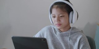 一个亚洲女孩晚上戴着耳机在平板电脑上通过网络学习。亚洲女孩的在线学习理念是戴着耳机在平板电脑上通过互联网进行在线学习。在家在线学习的概念