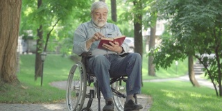 残疾人老人坐在轮椅上看书。广角照片积极的白人男性病人享受户外爱好在阳光明媚的夏天。老龄化和生活方式的概念。