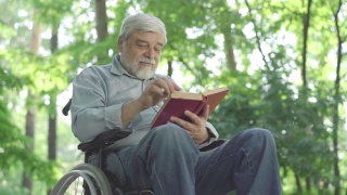 下角视角，吸收残疾人老人看书和微笑。聪明的残疾人白种退休人员在阳光明媚的夏日公园里与文学一起休息。视频素材模板下载